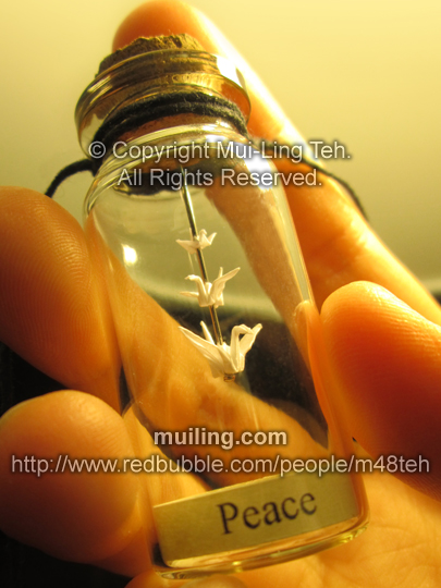 Three miniature origami cranes by Mui-Ling Teh, hanging on a needle in a bottle; with a yellow label on the bottom, with the word "Peace"