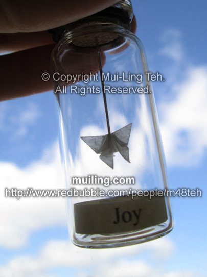 Miniature translucent white origami butterfly by Mui-Ling Teh, hanging on a needle in a bottle; with the word "Joy"  on a yellow label below.