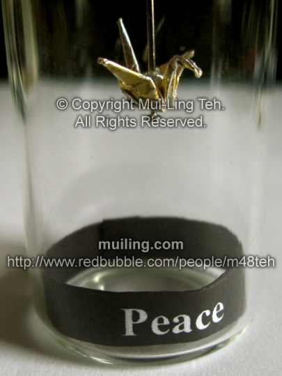 Miniature gold origami crane by Mui-Ling Teh, hanging on a needle in a bottle, with a black label on the bottom, with the word "Peace" written in bold white font.