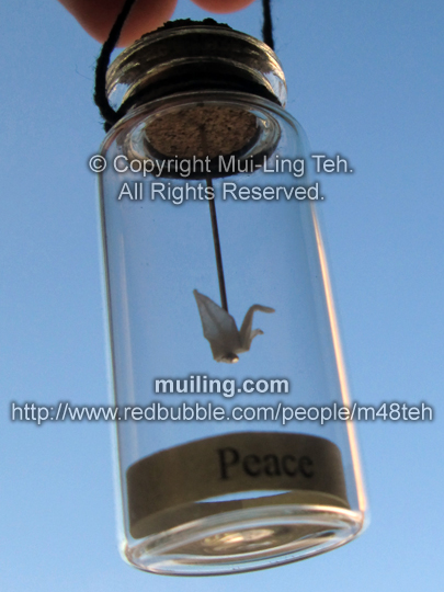 Miniature translucent white origami crane by Mui-Ling Teh, hanging on a needle in a bottle; with a yellow label on the bottom, with the word "Peace" written in black.