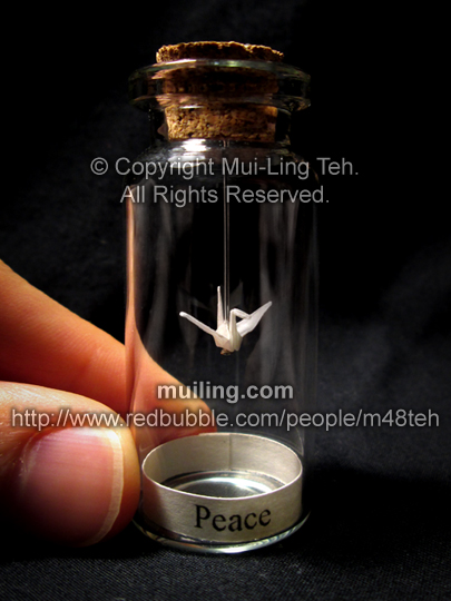 Miniature translucent white origami crane by Mui-Ling Teh, hanging on a needle in a bottle; with a yellow label on the bottom, with the word "Peace" written in black.
