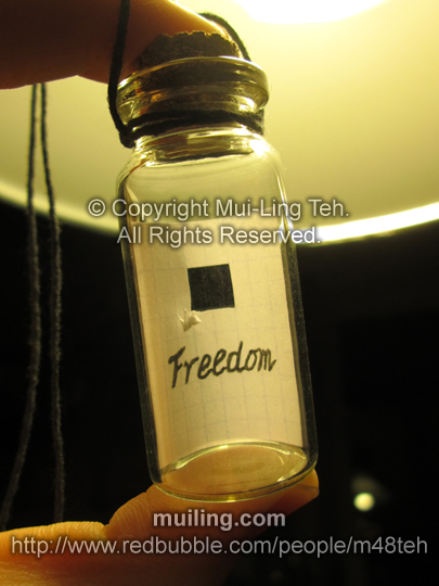 Miniature white origami crane in a bottle by Mui-Ling Teh, hanging off a piece of paper with the word "Freedom" written on it.