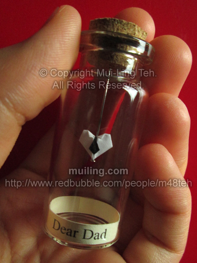 Miniature white origami heart with a black necktie by Mui-Ling Teh, hanging on a needle in a bottle. At the bottom is a yellow label with the words "Dear Dad" written in black.