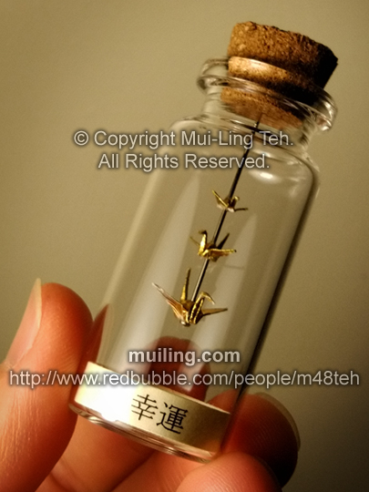 3 gold miniature origami cranes by Mui-Ling Teh, hanging on a needle in a bottle; with a yellow label on the bottom, with the word "good luck" written in Japanese