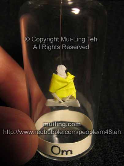 Miniature white origami meditation figure, wearing a yellow robe in a bottle, with 'Om' on a yellow label