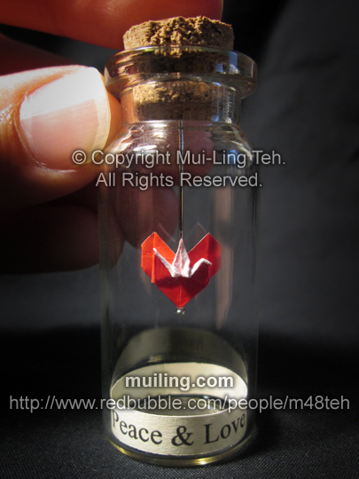 Miniture red origami crane and heart in a small bottle by Mui-Ling Teh, with the message Peace & Love written on a yellow label. The crane and the heart are made out of one paper.