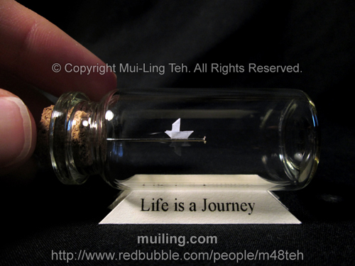 Miniature origami boat in a bottle by Mui-Ling Teh on top of a yellow base with "Life is a Journey" written on it.