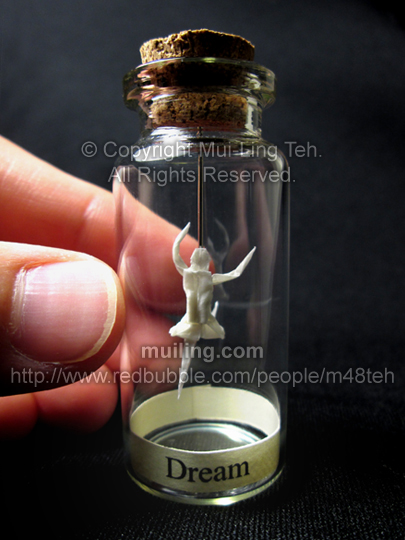 Miniature white origami ballerina by Mui-Ling Teh, hanging on a needle in a bottle; with the word "Dream"  on a yellow label below.